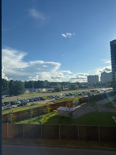 The image depicts a residential area in Vatutinki, Russia, on a sunny day. The perspective is from a high vantage point, likely a balcony or window, looking out over a street lined with parked cars. The scene is dominated by a bright blue sky with scattered white clouds.
In the foreground, a dark tiled surface suggests a balcony or rooftop. Beyond this, a road curves through the scene, with a long line of parked cars on the left side. The cars are of various colors, including black, white, and blue. To the right of the road, a grassy area is bordered by a wooden fence and a metal grate structure.
In the mid-ground, more buildings are visible, including multi-story apartment blocks. Trees and greenery are interspersed throughout the landscape, suggesting a well-developed but still somewhat natural environment. The sunlight casts long shadows, indicating it is likely morning or afternoon. The overall atmosphere is peaceful and ordinary, depicting a typical urban residential scene. There is no visible text in the image.