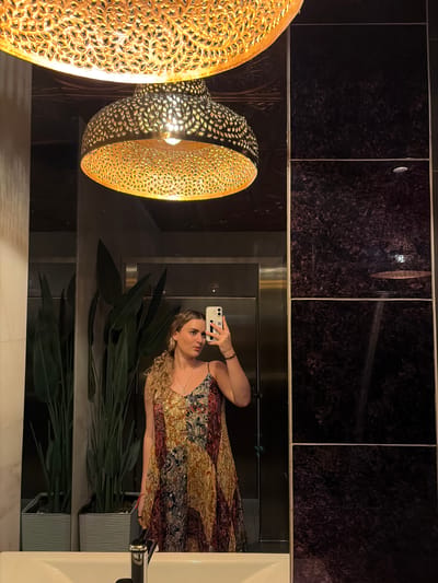 A woman is taking a selfie in a bathroom. The bathroom has dark granite tiles on the walls. Two decorative gold lamps hang on the left side of the frame. There are green plants, a white sink, and chrome hardware also visible. The location might be in Coral Gables, United States of America, but the image alone doesn't provide enough information to definitively confirm this.