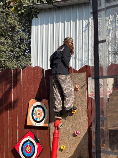 An individual with long brown hair, wearing a camouflage bandana, black long-sleeved shirt, and camouflage pants, is depicted in the process of ascending a vertical climbing structure. The person's left foot rests on a yellow climbing hold, while their body is partially turned, and they are looking over their right shoulder towards the camera.
The climbing structure consists of a panel of light-colored OSB or particle board affixed vertically to a reddish-brown wooden fence. Multiple colorful climbing holds are attached to this panel, including yellow, pink, and light beige. To the left of the climbing wall, two "Geologic soft archery" targets are mounted on the wooden fence: an orange rectangular target and a red square target, both featuring concentric rings and numerical scores. Below these targets and the climbing wall, a red vertical pipe, resembling a fire hydrant with a silver top, is present.
In the background, behind the wooden fence, a light-colored corrugated metal building is visible. To the left of this building, green trees and foliage are prominent under natural sunlight, which casts shadows across the scene. A partial view of a dark-framed window or transparent barrier is on the far right, behind which a white sheet or poster displaying Hebrew text is discernible.
The scene is located in the Menashe Regional Council, Israel.