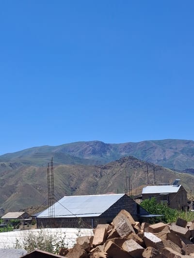 The image depicts a rural landscape with a clear blue sky. In the foreground, a pile of large, rough-hewn stones is visible. Behind the stones, two buildings with metal roofs are present. The building on the left is a long, rectangular structure with stone block walls and a silver metal roof. A tall, metal scaffolding structure stands next to it. The building on the right is a two-story house constructed from stone, with a silver metal roof and a solar panel on top. Green foliage surrounds the right building. In the background, rolling hills and mountains are visible, with layered vegetation and rocky outcrops. The scene appears to be a residential or agricultural area in Yeghegnadzor, Armenia, on a bright, sunny day. There are no people or animals visible in the image.