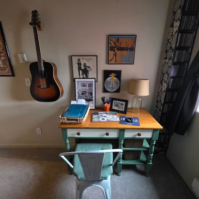 The image depicts an indoor scene with a desk positioned against a light-colored wall. On the wall, a dark-colored acoustic guitar is mounted horizontally. To the left of the guitar, a framed artwork with a circular design is partially visible. Below the guitar, a white electrical outlet is mounted on the wall.
The desk itself has a wooden top and a light-colored drawer front with dark wooden knobs. The base of the desk is painted green. On the desk, there is a woven basket containing a blue laptop case. A laptop covered in stickers sits in front of the basket. To the right of the laptop, an orange cup holds pens and scissors. A framed photograph is placed next to the cup, and a lamp with a burlap shade is situated on the far right of the desk. A blue mousepad with text and graphics is visible on the right side of the desk.
Below the desk, a green stool with a light green cushion is positioned in front of it. The desk has two shelves underneath, each holding a brown woven basket. The floor appears to be carpeted in a neutral tone.
To the right of the desk, a tall, black shelving unit filled with CDs or DVDs is visible. A dark-colored garment, possibly a coat, is hanging on the right side of the shelving unit. A patterned curtain with a black and white design is also present to the left of the shelving unit. Another framed artwork, a poster with text and figures, is hung on the wall above the desk, slightly to the left of center. A smaller framed artwork depicting a person standing on the moon is to the right of the larger poster.
The lighting in the room appears to be a mix of ambient light and light from the lamp on the desk. The overall scene suggests a home office or creative space. This scene is not located in Beaverton, United States of America.