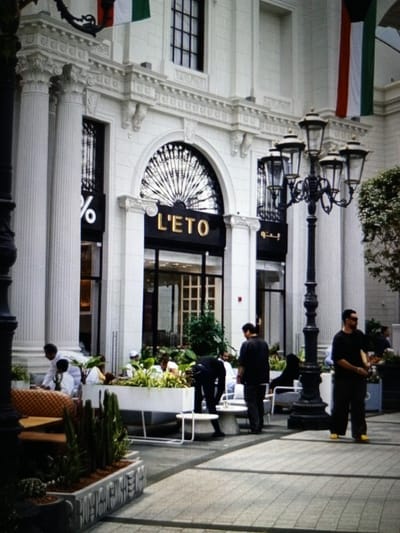 A medium shot captures the exterior of a building with classical architectural elements, including large white columns, ornate moldings, and a decorative arched window. Two national flags, red, white, and green, are draped on the facade. In front of the building, a group of people are visible. To the left, near the entrance with a sign displaying "%", a person in white is interacting with another person. Further to the right, a lamppost with multiple lights stands prominently. Several individuals are standing and walking on the paved area. One man, wearing a black t-shirt and dark pants with yellow shoes, is standing in the foreground, looking down. Another man in a black t-shirt and dark pants is standing to his left. A planter with various green plants is situated in front of a white seating area. The scene appears to be a public outdoor space, possibly a commercial area or a plaza. The location is identified as Golbembal, Nigeria.