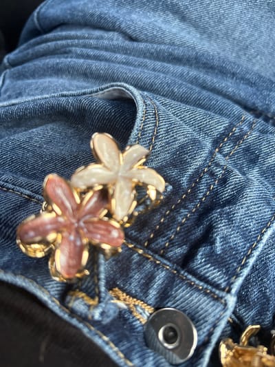 The scene depicts a close-up view of two decorative flower-shaped objects resting on blue denim fabric in Moțăței, Romania. The objects consist of two five-petal flower motifs, each approximately 2-3 cm in diameter, encased in gold-toned metal settings. One flower features off-white, translucent petals, positioned slightly above and to the right of a second flower displaying reddish-brown, translucent petals. A small gold-toned link is visible between the two flowers, suggesting they are connected or part of a single accessory. The underlying blue denim fabric exhibits a distinct twill weave, with prominent yellow-orange contrast stitching defining seams and a pocket edge. Multiple folds and creases are present in the denim. A circular, silver-toned metal button or rivet is partially visible in the lower right portion of the frame. No individuals or overt actions are discernible.