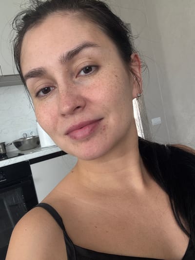 A woman with dark hair pulled back and a black strap visible on her shoulder is pictured from the chest up, looking directly at the camera. She has light skin with freckles and appears to be in a kitchen setting, with kitchen counters and appliances visible in the background. The lighting is natural, coming from the right side of the frame.