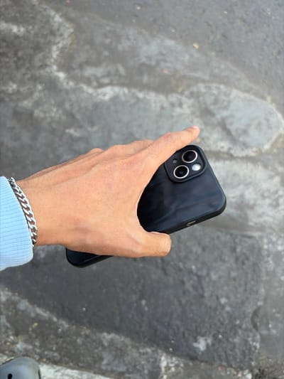 An individual's left hand, adorned with a silver chain bracelet and a light blue sweater sleeve, holds a black smartphone with a textured back. The phone has a distinctive camera module with two prominent lenses. The background is a textured, gray, asphalt-like surface, possibly a road or sidewalk, with some lighter, possibly wet, patches. The scene is outdoors, and the lighting suggests daytime. There is no specific identifiable landmark or context within the image to place it in Pekalongan, Indonesia.