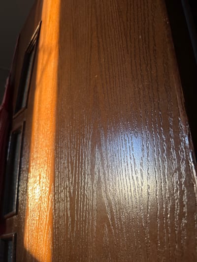 The image displays a close-up, angled view of a brown wooden or wood-laminate door in Mu'adammiyat ash-Sham, Syria. A strong, golden-orange band of sunlight illuminates the left third of the door, highlighting its vertical wood grain texture. Several dark, rectangular recessed panels or window inserts are partially visible along the extreme left edge of the door. The main panel of the door, extending to the right, exhibits the same brown color and vertical grain, with subtle reflections of light visible on its surface. No individuals or specific actions are depicted. A faint dark reddish fabric or object is discernible on the far left margin, adjacent to the door's edge.