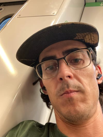 A close-up, low-angle, selfie-style photograph taken indoors, likely within a train carriage. The subject, a person with fair skin, is wearing a camouflage baseball cap, eyeglasses with thin metal frames, and a green t-shirt. They have dark hair and appear to be wearing wireless earbuds in their right ear. The background consists of a light-colored, smooth, curved wall, possibly part of the train's interior, with a circular window visible on the left. The lighting suggests an interior setting, with a strong light source reflecting off the wall. The person's gaze is directed towards the camera. There is no indication of the location being in Sant Cugat del Vallès, Spain.
