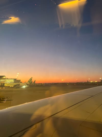 The image shows an airplane wing viewed from inside the cabin. Through the window, one can see the airport apron at Paray-Vieille-Poste, France, with other aircraft parked at gates. The sky transitions from blue to orange, indicating either sunrise or sunset. A Transavia aircraft is visible at one of the gates. Airport lighting is visible along the runway and around the apron. The scene suggests the airplane is either preparing for takeoff or has recently landed.