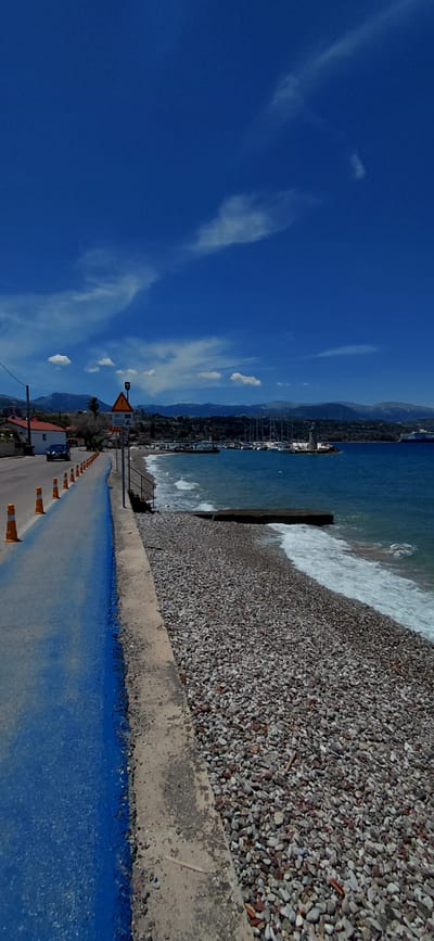 A bright, sunny day is captured in this photograph of a coastal road and beach in the Municipal Unit of Aegio, Greece. The sky is a vibrant, cloud-dotted blue, with distant mountains forming a hazy backdrop. On the left, a paved road with a distinct blue painted lane runs alongside a rocky beach. Orange traffic cones line the edge of the road, and a car is visible in the distance. A signpost with several warning symbols stands near the road. The beach itself is covered in smooth, grey and brown pebbles, with waves gently lapping at the shore. In the distance, a harbor with numerous sailboats and a lighthouse can be seen. A large ship is also visible on the horizon. The scene appears peaceful and idyllic, with no people or animals present.