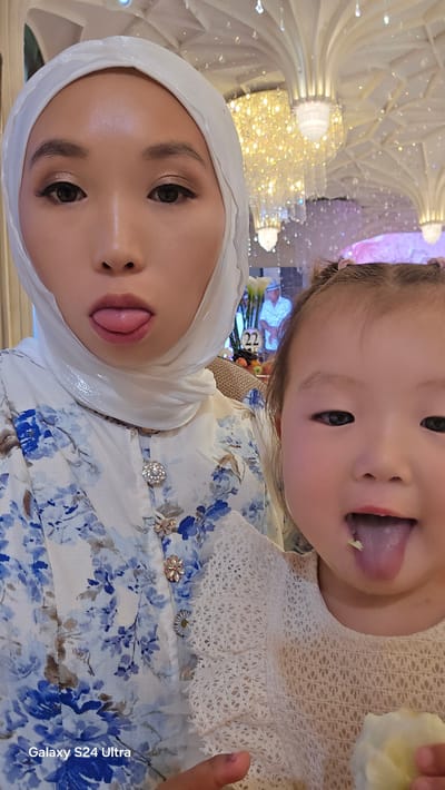 The image shows a close-up, selfie-style shot of two people, likely a mother and child, posing for a photo. The older subject, wearing a white hijab and a floral dress with blue and white patterns, sticks out her tongue playfully. She has noticeable makeup, including eyeshadow and defined eyebrows. The younger subject, a child, is in the foreground and also sticks out her tongue, with a bit of light-colored food on it. She is wearing a white lace garment. Both individuals appear to be indoors, with a brightly lit, ornate ceiling and chandeliers visible in the background, suggesting a festive or formal indoor venue, possibly a restaurant or event space. The overall mood conveyed is lighthearted and fun. The image also contains the text "Galaxy S24 Ultra" in the top left corner.