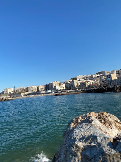 The image depicts a coastal view in Beau Fraisier, Algeria. The scene features a clear, cloudless blue sky dominating the left side of the frame. The right side showcases the Mediterranean Sea with water ripples and a large rock formation in the foreground. Adjacent to the sea, a dense row of buildings lines the coast, appearing as apartment blocks or commercial structures. A paved walkway runs parallel to the buildings along the coastline. The overall environment suggests a bright, sunny day in a densely populated coastal area.