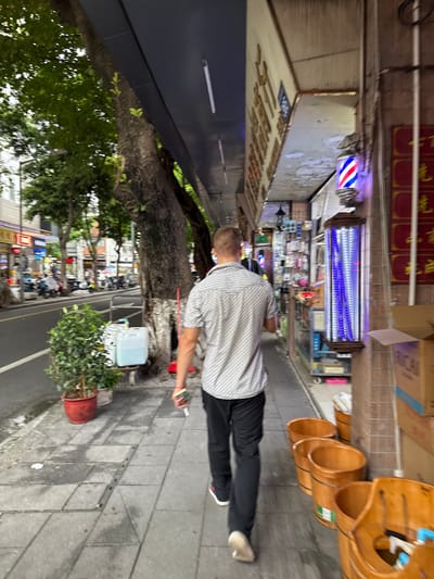 A man with short, blond hair walks away from the camera down a sidewalk in Liwan District, China. He is wearing a white and gray patterned short-sleeved button-up shirt and black pants. He is holding a small, light-colored object in his right hand. To his left, a tree with rough bark grows alongside the sidewalk. The sidewalk is paved with gray tiles and is wet in some areas. To the left of the sidewalk, a street is lined with parked motorcycles and scooters. Buildings with awnings and signs line the right side of the sidewalk. One building has a barber pole with red, white, and blue stripes illuminated with purple light. Several large, wooden buckets are placed on the sidewalk near the buildings. The sky is overcast and the lighting suggests it is daytime.