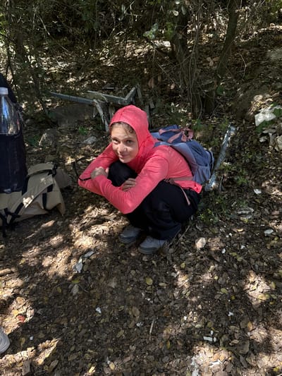 An overhead photograph captures a young female individual pausing in a dry, wooded environment within the Mate Yehuda Regional Council, Israel. The person is sitting or crouching on the ground, which is covered with an abundance of brown, dry leaves, dirt, small rocks, and patches of green vegetation. She is dressed in a bright coral-pink long-sleeved hooded top, black pants, and grey athletic or hiking shoes. A blue and pink backpack is worn on her back, and a tan-colored bag or backpack rests on the ground near her. A clear water bottle is also visible nearby.
She looks directly up towards the camera with a calm, neutral expression. The setting features dappled sunlight filtering through trees, casting distinct shadows on the ground, indicating a bright, sunny daytime. In the background and mixed with the leaf litter, there are scattered pieces of white debris, possibly plastic or other litter, and some metallic-looking fragments. The scene suggests a break during an outdoor activity like hiking or exploring. There is no visible text.