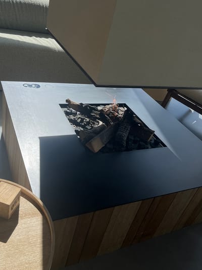 The scene is an interior or semi-exterior lounge space located in Overveen, Netherlands. A rectangular fire pit serves as the central subject. It features a dark, matte grey top surface, likely metal, with a central square aperture. Within this aperture, a gas-fueled fire exhibits small orange flames among a composition of natural and artificial logs and dark rock fragments. Faint circular indentations and a dark logo are visible on the fire pit's top surface to the left. The fire pit's base consists of diagonally oriented light-brown wooden planks. In the lower left quadrant, a segment of a circular, light-toned wooden side table is visible, upon which rests a wooden block marked "051". The upper background reveals a light grey upholstered seating structure and a light-colored wall. Strong natural light, originating from the left, illuminates the scene, casting sharp shadows on surfaces including the fire pit, seating, and side table. The floor is dark grey. No human subjects are present.