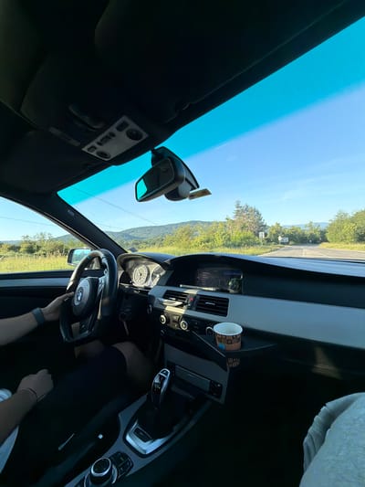 The image depicts the interior of a car, with a person's hands visible on the steering wheel. The view looks forward through the windshield, showing a road winding through a landscape of rolling hills and trees under a clear blue sky. The dashboard of the car is visible, including the steering wheel, gear shift, and a cup holder with a paper cup. The car appears to be in motion on a road outside of the city of Хаджийско, Bulgaria, given the presence of natural scenery and the absence of urban structures. The time of day appears to be late afternoon or early evening, as indicated by the sunlight casting shadows and illuminating the landscape.