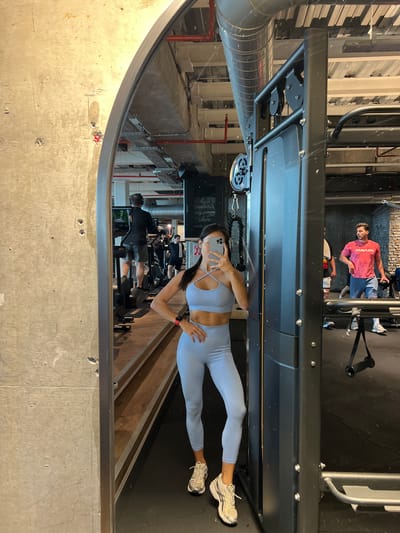 A woman is taking a mirror selfie in a gym. She is wearing a light blue sports bra and leggings. The gym has exposed ceilings with pipes and lighting fixtures, concrete walls, and wooden floors. In the reflection, other gym-goers can be seen using exercise equipment. One man in a red t-shirt and blue shorts is standing near weight racks, and another man is on a treadmill. The woman is holding her phone up to take the photo, and her left hand is on her hip. The gym appears to be a modern, industrial-style space.