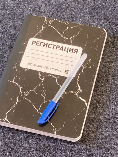 A blue and clear ballpoint pen rests diagonally across a black and white marbled notebook. The notebook is open, with its cover facing upward on a dark, textured surface, possibly carpet or fabric. The notebook's cover features a label with Cyrillic text that reads "РЕГИСТРАЦИЯ" and "100 листов • 200 страниц". The pen is positioned with its blue cap and clip on the lower left side and its clear barrel extending towards the upper right. The background is a dark, grey, and fibrous material, likely a floor covering. The lighting suggests an indoor setting. There is no indication of the location being Arbagish, Uzbekistan.