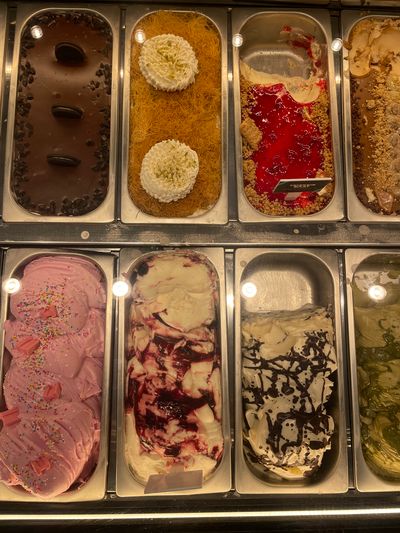 The image displays a top-down view of a gelato display case filled with various flavors of gelato. The display case is divided into six rectangular compartments, arranged in two rows of three.

In the top row, from left to right:
*   The first compartment contains a dark chocolate gelato topped with what appear to be chocolate chips and three whole Oreo cookies.
*   The second compartment features a bright orange, shredded dessert base, possibly kadaif, topped with two dollops of white cream, garnished with green sprinkles.
*   The third compartment has a mostly empty metal tray with remnants of a creamy yellow gelato, overlaid with a thick, vibrant red sauce and a scattering of crushed cookie or nut pieces. A small rectangular label is visible on the right side of this compartment.

In the bottom row, from left to right:
*   The first compartment is filled with a vibrant pink gelato, generously sprinkled with colorful sprinkles and topped with several small, rectangular pink candy pieces.
*   The second compartment contains a white gelato swirled with streaks of a dark red sauce, likely a fruit compote.
*   The third compartment holds a white gelato with a dark chocolate sauce drizzled in a marbled pattern.
*   The partially visible compartment to the far right of the bottom row appears to contain a green-colored gelato.

The gelato display is illuminated by overhead lights, which are reflected in the metallic surfaces of the display trays and the glass covering. The overall impression is of a well-stocked dessert shop. Based on the visible label in the third compartment of the top row, which reads "TZTZEIK ΠΛΑΤ" and appears to be in Greek, the location is likely in Thessaloniki, Greece, as this is a common transliteration and the presence of Greek text strongly suggests this region. The "Municipal Unit of Thessaloniki" is the relevant administrative division.