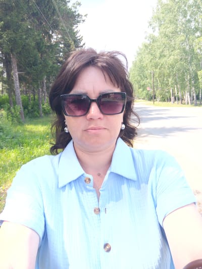 A woman with dark brown hair and dark sunglasses is taking a selfie outdoors. She is wearing a light blue collared shirt with three visible buttons. She has on spherical earrings. Behind her is a paved road with trees lining both sides. The trees on the left appear to be coniferous, while those on the right are deciduous, with light-colored bark. The ground on the left side of the road is covered in green grass and some yellow wildflowers. The sky is bright and appears to be partly cloudy. The overall impression is a casual outdoor scene, likely during daytime in a rural or semi-rural area. The location is Zalesovo, Russia.