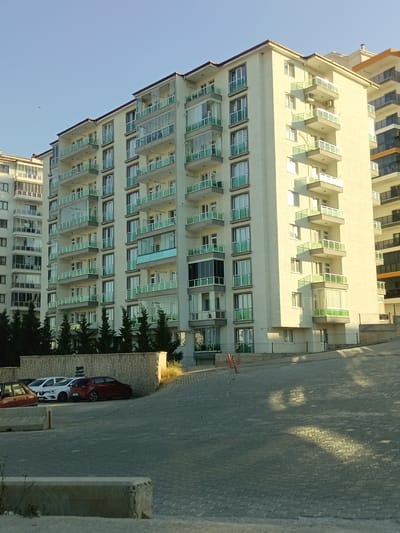 The image shows a modern apartment building in Yahşihan, Turkey. The building is tall and cream-colored, with multiple balconies featuring green glass railings. Several other apartment buildings of varying heights are visible behind and to the left of the main building, some with darker facades and balconies. The sky is clear and blue. In the foreground, a paved area with parking spaces is visible. A few cars are parked, including a red sedan and a white hatchback. A low, textured stone wall runs along the parking area, behind which dark green trees are planted. The scene appears to be in a residential area, likely an urban or suburban setting.