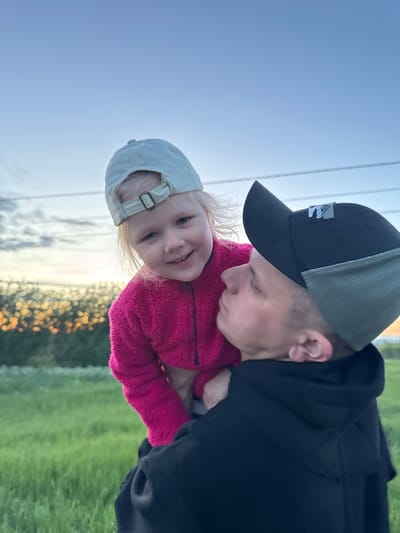 A man and a young child are depicted in a close-up shot. The man is wearing a black and olive green baseball cap with a small logo on the front. He has short, light-colored hair. He is wearing a black hoodie. The child is wearing a light gray baseball cap and a bright pink fuzzy jacket. The background is a mix of sky, blurred vegetation, and what appears to be an outdoor setting. The precise location in Горный, Russia, cannot be determined from the image.