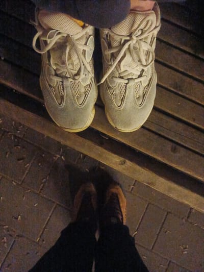 The image shows a close-up, overhead view of two pairs of feet wearing athletic shoes. The top pair of feet, partially obscured by dark fabric, are wearing light-colored sneakers with visible lacing and mesh detailing. They are positioned on a slatted wooden surface. The bottom pair of feet, also partially obscured, are wearing darker shoes and are standing on a paved surface that appears to be cobblestone or brick. The overall lighting suggests it is nighttime or a dimly lit indoor environment. There is no information in the image to suggest a specific location in the city of Radkevshchina, Russia.