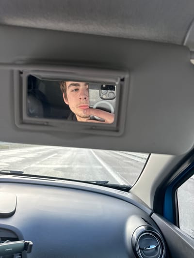 The image displays the interior of a vehicle, with the perspective directed towards the front windshield. The primary subject is the reflection of a male individual in the passenger-side vanity mirror, which is flipped down. This individual has light brown hair and facial stubble, and is seen holding a smartphone with his right hand, positioned to take a selfie. The rear of the smartphone, featuring multiple camera lenses and a light-colored case (possibly with a MagSafe ring), is visible in the reflection. Below the mirror, the vehicle's dark, textured dashboard is visible, along with a portion of the windshield wipers at its base. A circular passenger-side air vent and a phone mount on the far left of the dashboard are also discernable. Through the windshield, an asphalt road with distinct white lane markings stretches forward, appearing bright and reflective. There are no identifiable landmarks or signage within the image to confirm the specific location within Ribeira de Pena, Portugal.