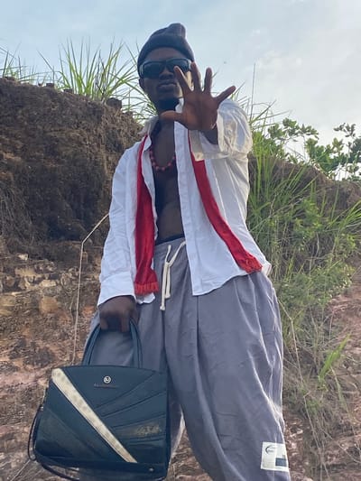 This image is a full-body outdoor portrait of a man, likely in Afikpo, Nigeria, as per the provided context. The man, with dark skin, is centered in the frame, looking directly at the viewer. He wears a black beanie, dark sunglasses, and a red and black beaded necklace. His clothing includes a white long-sleeved shirt with prominent red fabric details on the sleeves and a vertical red stripe down the front, worn over a dark undershirt or with his upper chest exposed. He is also dressed in loose-fitting, light grey trousers.
The man's left hand is raised, making a peace sign gesture, while his right hand holds a black shoulder bag. The bag features a diagonal light-colored stripe across its front and a circular logo containing a stylized letter, possibly a "B" or similar design. A small white label with an unreadable black symbol is visible on the upper right side of his grey trousers.
The setting appears to be a natural, uncultivated area. The ground consists of earthy, rocky terrain with patches of wild green grass and some leafy foliage in the background, suggesting a hillside or an undeveloped outdoor location. The sky is bright and appears overcast, indicating daytime. The man's pose and direct gaze convey a sense of casual confidence.