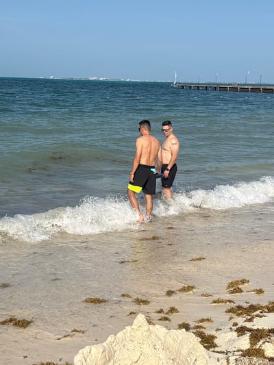 This media file depicts two men standing in shallow water at a beach. One man is closer to the camera, facing away from it, while the other is slightly further back and turned more towards the camera. Both are wearing swim trunks. The man closer to the camera has black swim trunks with a yellow stripe. The man further back has black swim trunks and is wearing sunglasses. The scene is set at a beach with calm, clear water lapping at the shore. The sand appears light in color and is partially covered with seaweed. In the background, a long pier with lights stretches out into the ocean, and a distant shoreline with buildings can be seen. The sky is a clear, bright blue, suggesting it is daytime with fair weather. The overall mood appears relaxed and casual, consistent with a beach outing. The location is identified as Punta Sam, Mexico, which aligns with the visual cues of a tropical beach setting and a pier common in such areas. No visible text is present in the image.