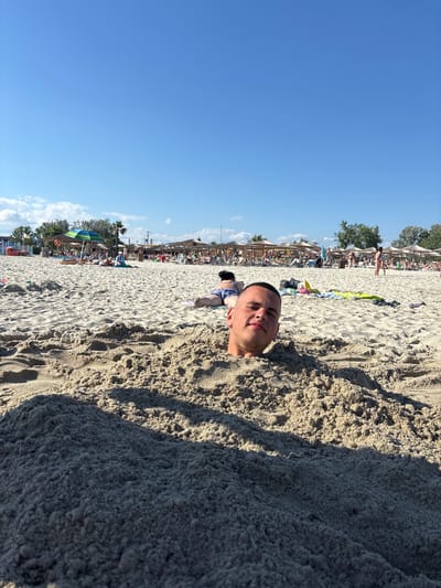 A young man is buried in the sand on a sunny beach. His head and shoulders are visible above the sand, and he is looking towards the camera with a slight smile. In the background, there are other beachgoers, umbrellas, and lounge chairs. The sky is clear and blue. The scene appears to be a popular tourist destination.