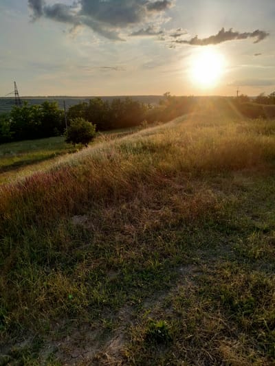 This is a photograph taken in Ananiv, Ukraine, during a sunset. The image captures a rural landscape with rolling hills covered in tall grass and sparse trees. The sun is setting on the horizon, casting a warm, golden light across the scene, creating a lens flare that streaks across the frame. In the distance, power lines and a few scattered buildings are visible, suggesting a rural or semi-rural setting. The foreground is dominated by a grassy slope, with a narrow dirt path winding through it. The sky is partly cloudy, with some darker clouds contrasting with the bright sunset. The overall mood of the image is serene and peaceful, evoking the beauty of a Ukrainian sunset in the countryside.