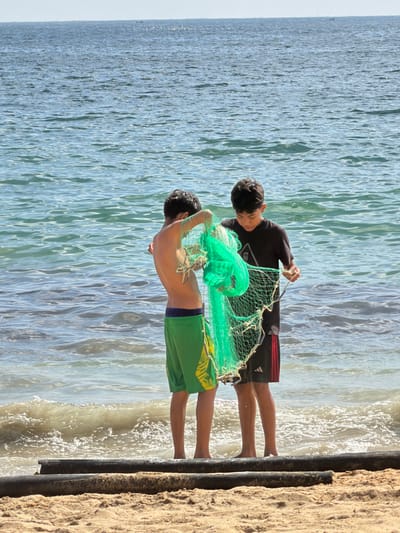 Two people are on a beach holding up a fishing net. The person in the foreground is shirtless and wearing green shorts with a blue stripe. The person behind is wearing a black shirt and black shorts with a red stripe. The fishing net is green and appears to be tangled. The water is on the left, and the sand is on the right. A metal barrier separates the sand and the water.