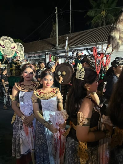 The image depicts a nighttime outdoor event, likely a procession or cultural performance, in Munggu, Indonesia. Three women in traditional Balinese ceremonial attire are positioned in the foreground. The two women on the left face the camera, smiling, wearing elaborate gold-colored headpieces, ornate red and gold necklaces, gold armbands, and patterned sarongs. They have white sashes with red markings and hold bundles of crumpled white and red-stained cloth. The woman on the right is seen in profile, wearing similar attire with a decorative hair comb and a prominent gold ear ornament.
In the midground and background, numerous other individuals, predominantly men in traditional Balinese dress including head wraps, are participating in the event. On the far left, a large, multi-tiered white and gold ceremonial float or structure is visible, with people seated or standing on it. To the right, a man is seen striking a large, circular gong with a mallet. Another ceremonial structure featuring prominent red beaded strands and a large, dark fur or feathered plume is also present.
The background includes buildings with dark tiled roofs, a visible power line system on poles, and a palm tree silhouetted against the dark night sky. A store sign is partially visible, bearing the text "AFDAN GORDE." Artificial lighting illuminates the scene, casting shadows, particularly on the faces of the foreground women.