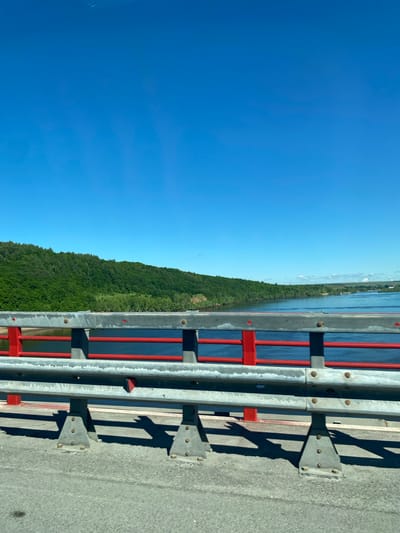 The image shows a roadside view from a bridge on a clear, sunny day, with the camera positioned looking along the bridge railing. The railing is gray with red vertical supports. Beyond the railing is a body of water and a heavily forested shoreline. The sky is a clear, bright blue. The location is likely near Sovkhoza "Pyatiletka", Russia, as requested.