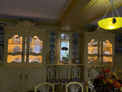 The image depicts the dining room of Claude Monet's house in Giverny, France. The room features yellow-painted walls, ceiling, and wooden furnishings. Two tall, light-yellow display cabinets with ornate tops and glass doors are centrally positioned, internally illuminated to showcase numerous blue and white ceramic plates, bowls, and teacups. Between these cabinets, and to the right, several blue and white decorative plates are mounted vertically on the wall. A large blue and white lidded vase rests on a shelf below the wall-mounted plates. A bright yellow, bowl-shaped glass chandelier with a dark accent hangs from the ceiling. In the foreground, several light-yellow wooden chairs with spindle backs and patterned seat cushions are partially visible, positioned around an unseen dining table. A large bouquet of colorful flowers, predominantly red, orange, and yellow dahlias, along with bare branches, occupies the lower right corner of the frame. A window with white sheer curtains is visible in the background, revealing outdoor greenery. Portions of framed artwork are discernible on the far left and right walls. No individuals are present.