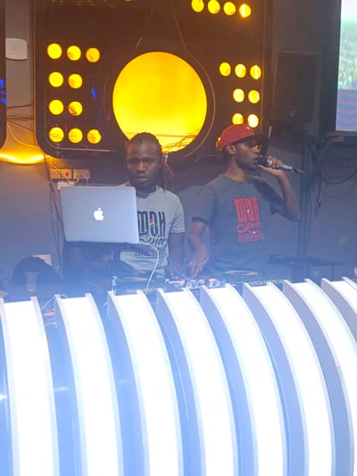 In Syokimau-Mulolongo ward, Kenya, two men are working as DJs. The man on the left has dreadlocks and is wearing a grey t-shirt. He stands behind a laptop with an Apple logo. The man on the right is wearing a red cap and a grey t-shirt. He is holding a microphone and speaking into it. Both men are behind a DJ console. Behind them is a light fixture with a large circular yellow light and smaller circular lights around it. A speaker is visible to the right.