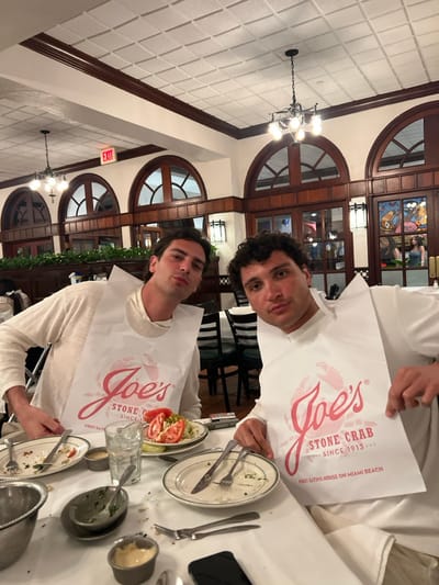 The image shows two young men seated at a table in a restaurant located in Miami Beach, Florida, at the coordinates provided. The restaurant is Joe's Stone Crab, the first eating house on Miami Beach, and the two men are both wearing white bibs with the restaurant's logo on them. The men are seated across from each other, and both have their hands on the table. There are a few empty plates, silverware, and a glass of water on the table in front of them. The restaurant has a tiled ceiling, wooden trim, and large windows. The environment is well-lit and clean. The men appear to be enjoying their meal. The image was taken indoors during the day.