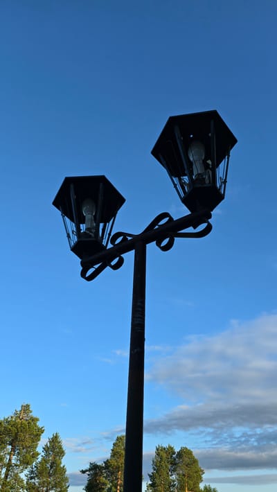 A low-angle, full shot shows two black, hexagonal streetlights on a dark, textured pole. The streetlights are silhouetted against a clear, deep blue sky with scattered, wispy white clouds. Each streetlight contains a visible light bulb. The pole extends from the bottom center of the frame upwards. At the bottom of the frame, the tops of several evergreen trees are visible, indicating an outdoor, possibly park or roadside, setting in Верхнеказымский, Russia.