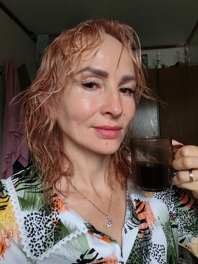 A close-up, selfie-style portrait shows a Caucasian woman with wet, reddish-blonde hair, wearing a brightly patterned shirt with green, orange, yellow, and black designs. She holds a dark glass mug of coffee in her right hand, which also displays a silver band on her ring finger. The woman's face is turned slightly to her right, and she has dark eyeliner and pink lipstick. She appears to be indoors, possibly in a bathroom or bedroom, with a wooden closet or cabinet visible in the background. Towels hang on a rack on the left side of the image. The scene suggests a casual, personal moment. The backdrop suggests the general environment of a home.
