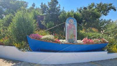 This image depicts an outdoor scene with a blue boat filled with flowers. The boat is positioned on a white, tiered structure. A mosaic of a religious figure holding a palm branch is affixed to the front of the boat. Behind the boat, there are various trees and bushes. To the left of the boat, a large dark rectangular image featuring a man in dark clothing is visible against the foliage. The sky is clear and blue. This scene is located at the Bilhorod-Dnistrovskyi Fortress in Bilhorod-Dnistrovskyi, Ukraine.