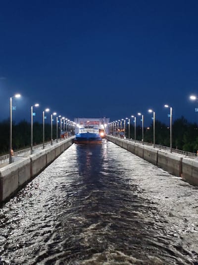 The image depicts a vertical shot of the Sheksna river lock in Sheksna, Russia, at twilight. A large, blue and white cargo ship is centrally positioned within the concrete lock. Both sides of the lock are lined with concrete walls and numerous lampposts, each casting light onto the water. The sky above is a deep, clear blue, indicating nighttime or very late dusk. The water in the lock is turbulent, with ripples and reflections of the lights. Trees are visible in the background on the right side of the image.