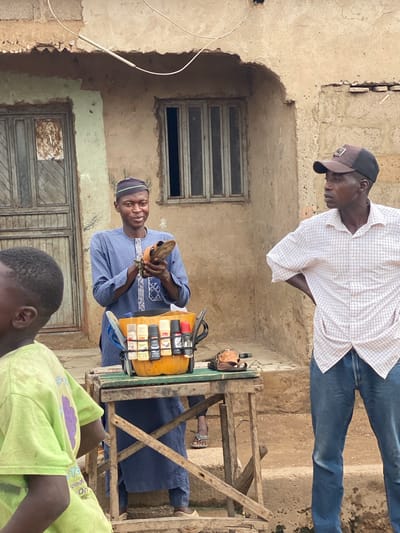 In Rigachikun, Nigeria, a man in a blue patterned robe and a dark kufi cap holds a brown sandal, examining it. He stands behind a wooden stand displaying a collection of shoe polish bottles in a yellow container. To his right, another man in a checkered long-sleeved shirt and blue jeans stands with his hands on his hips, wearing a baseball cap. In the foreground, a young person in a bright green t-shirt is partially visible. The background features a weathered building with a wooden door and a barred window. The ground appears to be dirt.