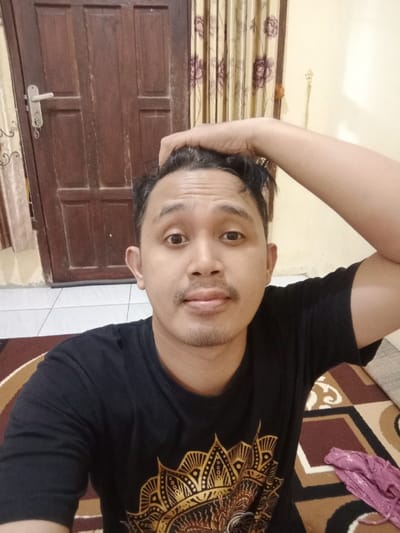 A male individual is depicted indoors in Brangsong, Indonesia. He is positioned facing forward, looking directly into the camera with a neutral expression. His left hand is placed on top of his head. He wears a short-sleeved black t-shirt featuring a prominent circular gold and brown patterned design on the chest. The background consists of a dark brown wooden door with a silver handle on the left, and a light-colored wall with a patterned curtain on the right. A decorative item is affixed to the wall next to the curtain. The floor features a brown, beige, and black geometric patterned rug, with white tiled flooring visible in the lower left portion of the image. A crumpled piece of pink/purple fabric rests on the rug in the lower right.