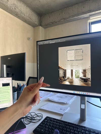 A woman's right hand, with long, manicured nails, is visible in the foreground, gesturing towards a computer monitor displaying architectural renderings. The monitor shows a project titled "RESIDENTIAL HOUSE LIVING ROOM 42.3 M2," featuring a floor plan and an interior visualization. The woman's arm extends from the left side of the frame, holding a mouse.

The computer setup includes a main monitor and a secondary laptop, with a keyboard and a mouse in front. Pens and papers are scattered on the desk. The environment appears to be an office with exposed concrete beams and a light-colored wall. The presence of a laptop, desk, and computer equipment suggests a workspace. There are no explicit visual cues within the image to pinpoint the location in Zovuni, Armenia; this detail is based on external information.