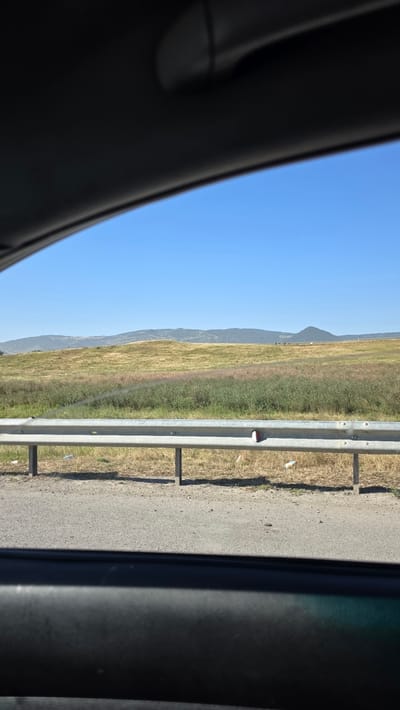 The image displays a roadside view of a grassy field under a clear blue sky. A silver guardrail runs horizontally across the lower portion of the frame, separating the asphalt road from the field. The field itself is a mix of dry, yellowish grass and greener, taller vegetation, with some areas appearing somewhat patchy. In the distance, rolling hills with sparse vegetation form the horizon. The scene suggests a rural or semi-rural environment. The foreground shows a dark interior surface, possibly the car's dashboard or window frame, indicating the photograph was taken from inside a vehicle. There are no visible people or specific landmarks that would pinpoint the location in Nizhniy Chiryurt, Russia, other than the general landscape being consistent with such a region. The lighting suggests it is daytime, likely mid-morning or afternoon, given the bright sunlight.
