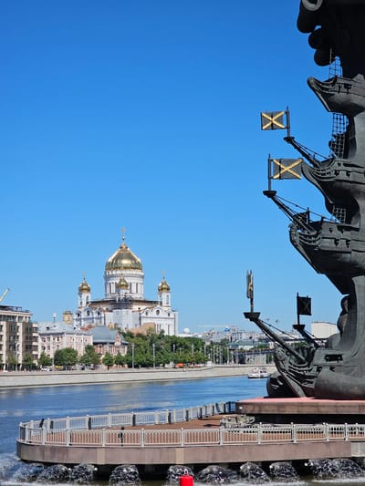 The image depicts a scene in Moscow, Russia, featuring the monument to Peter the Great and the Cathedral of Christ the Saviour. In the foreground, a large, dark bronze monument of a ship with multiple masts and flags is partially visible, dominating the right side of the frame. The ship's masts fly two black flags with golden "X" shapes. To the left of the monument, a circular waterfront promenade with white railings and wooden decking is prominent, with fountains spraying water into the river below. The Moscow River flows between the promenade and the far bank. On the far bank, the white Cathedral of Christ the Saviour, with its prominent golden dome and smaller golden domes, stands as a central landmark. Surrounding the cathedral are several other buildings, including apartment blocks and older architectural structures, interspersed with trees. The sky is a clear, vibrant blue. The overall scene is bathed in daylight, suggesting it is daytime. There are no people clearly visible in the frame. The location is identified as Moscow, Russia, specifically near the monument to Peter the Great and the Cathedral of Christ the Saviour.