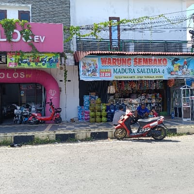 The image depicts a storefront in Kecamatan Brangsong, Indonesia (latitude: -6.953, longitude: 110.22) with a sign that reads "WARUNG SEMBAKO MADURA SAUDARA 4". The sign is blue and white and has the words "BUKA 24 JAM" in a smaller yellow square at the top left. There is a red motorcycle parked in front of the store. Two men and a child are sitting in front of the store. The men appear to be interacting with the child. There is a green, leafy canopy above the shop with wire strung through it. The storefront appears to be a convenience store selling various items. There is a pink sign with the words "LOS EV" in white letters. The storefront is located next to a sidewalk that is lined with black and white tiles. There are potted plants in front of the store. The storefront is located in an urban environment.