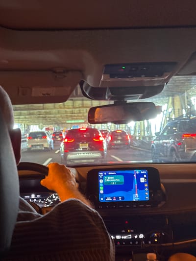 This media file depicts the interior of a vehicle, likely a car, with a view of a traffic jam outside. The scene is oriented vertically, suggesting it was captured with the phone held sideways.
In the foreground, a person's hands are visible, one of which is gripping the steering wheel. The person is wearing a cap. The dashboard is partially visible, featuring a GPS navigation device displaying a route on a map with traffic information. The time on the device reads "10:02" and later "10:32". The text on the GPS indicates "1H Hudson Pkwy", a highway in New Jersey, United States.
Outside the vehicle, a long line of cars is stuck in traffic. The brake lights of the cars are illuminated, indicating a standstill. The environment appears to be urban, with tall structures visible in the background, possibly buildings or overpasses. The lighting suggests it is daytime, but the specific weather conditions are not discernible. The overall mood is one of being stuck in traffic, with a focus on the driver's perspective and the navigation system.