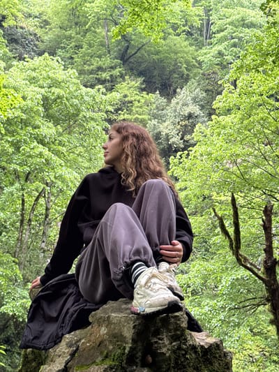 A young person with curly brown hair is perched on a moss-covered rock. They are wearing a black hoodie and gray sweatpants, with white sneakers and black and white striped socks visible. The person is looking down, appearing contemplative or observing something below. The rock is located amidst a lush green forest with trees filling the background. Based on the location information, the scene takes place in Tsabal, Abkhazia.