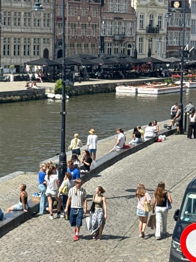 The image depicts a scene along the Leie River in Ghent, Belgium, on a sunny day. Several groups of people are visible along the cobblestone embankment and on the waterfront.
In the foreground, a man wearing a striped shirt and blue shorts walks towards the right. To his right, two women are walking away from the camera, one carrying shopping bags. To the left of the man, a group of young people are gathered, some sitting on a low wall, others standing. One young woman is sitting on the embankment to the far left, facing away from the camera.
Further in the background, more people are seen sitting at outdoor tables under umbrellas, suggesting a cafe or restaurant setting. Several boats are docked along the river, with one prominent boat displaying the text "Bordeaux Deward" twice. The architecture of the buildings lining the river is characteristic of Ghent, with ornate facades and multi-story structures. A lamppost stands prominently in the middle ground. The water of the river is calm and reflects the sunlight.